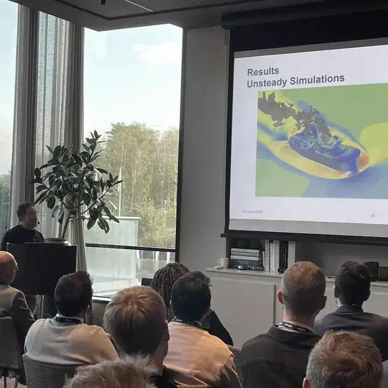 Audience seen from behind, a person standing at a lectern and a projected slide reading “Results” and “Unsteady Simulations” with a colour contour plot, in a room with tall windows
