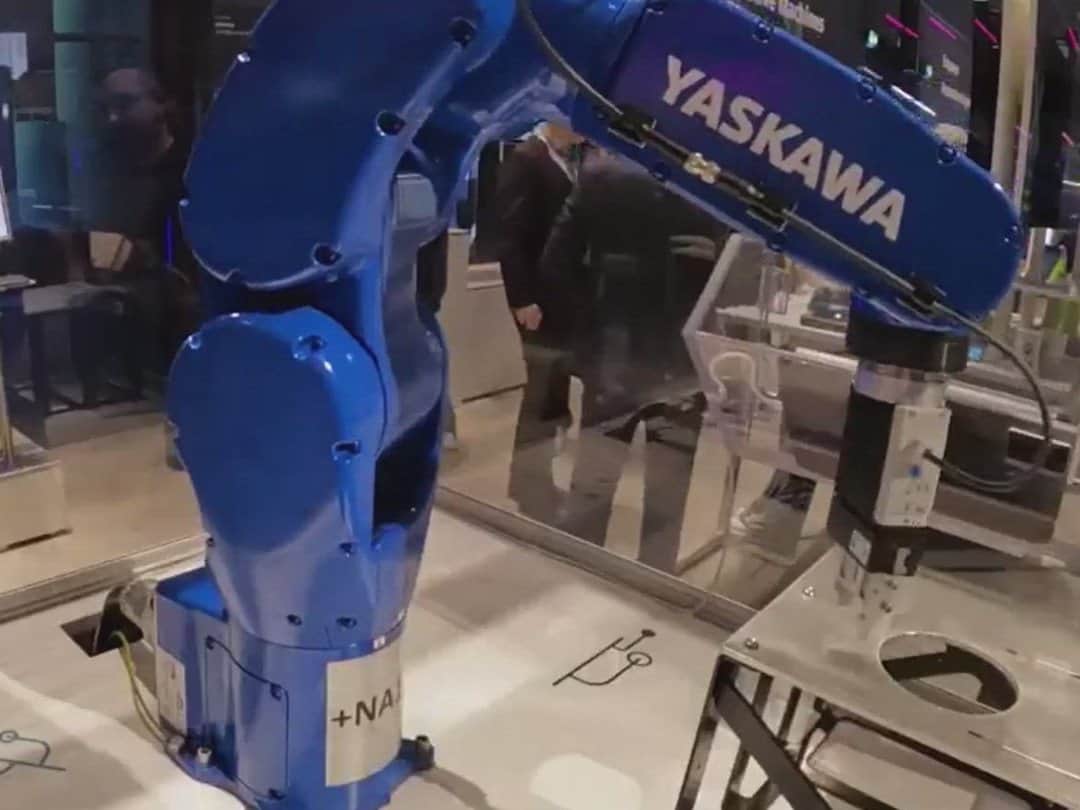 A blue industrial robot arm pressing on a metal test part in a glass-walled demonstration cell